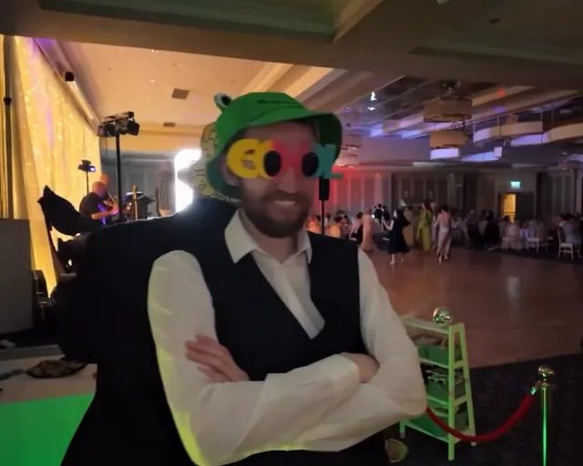 Guest in a green frog bucket hat and novelty glasses, arms folded